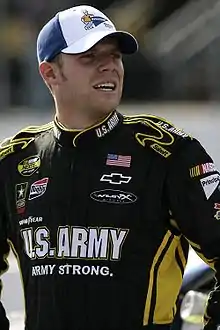 A man in his twenties wearing a white and blue baseball cap, and a black racing suit.