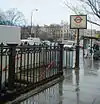 A white sign reading "REGENT'S PARK STATION BAKERLOO LINE" in black letters with people walking behind it all under a light blue sky