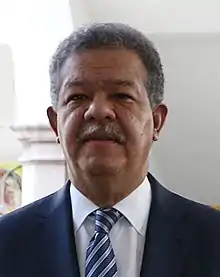 A portrait shot of a middle-aged man smiling somewhat and looking straight ahead. He has light brown skin, slightly African facial features, curly dark hair. He is mustachioed and wears a suit and tie.