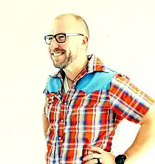 A balding Caucasoid man in glasses is pictured from the waist up; facing and looking to his right, his hands are on his hips, and he is wearing a brightly-colored, button-up collared shirt.