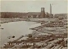 Sepia-tone photo of a pond surrounded by large logs. At the far end of the pond is a large building with a square tower and two smokestacks. Label is "T&T L CO MILL POND RICKETTS PA 1903" (i.e. Trexler and Turrell Lumber Company Mill Pond ...)