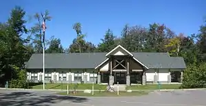 Photo of a modern one-story building with five windows and a two-story entrance with wooden framing and "Ricketts Glen" in metal letters. There are trees on three sides, with a grassy lawn and parking lot in front, and a bright blue sky.