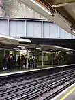 Eight people walking towards an escalator on a railway platform next to a green-tiled wall on the left and a railway track on the right