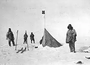 Four figures in heavy clothing stand near a pointed tent on which a small square flag is flying. The surrounding ground is ice-covered. Ski and ski poles are shown on the left.