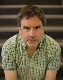 A brown haired man in a green plaid shirt looks into the camera while sitting on a ladder step.