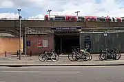 A grey building with a rectangular, dark blue sign reading "RUISLIP GARDENS STATION" in white letters all under a light blue sky with white clouds