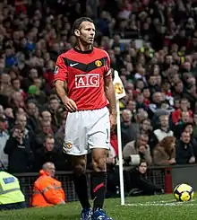 A white football player with short, dark, greying hair. He is wearing a red shirt, white shorts, white socks and white football boots. He is running and has puffed-out cheeks.