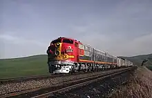 Red diesel locomotive with yellow striping leading silver train cars through rolling countryside