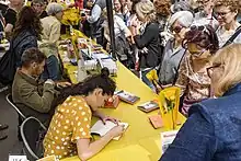 An author signs a book at a table seated by other authors and surrounded by a crowd.