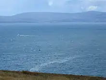 The attack site today, seen from a cliff above the bay. A small green wreck buoy is a few hundred metres away. A thin slick of oil is on the surface of the sea.