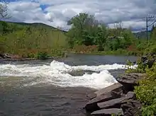 Whitewater forming where water joins a wide stream from the right. There are mountains and trees in the background, with some rocks at the lower right