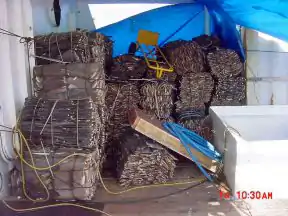 Small tied bales of flat gray objects, messily stacked underneath a translucent tent-like roof between white walls.