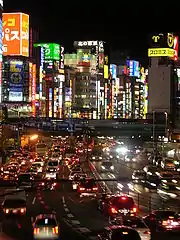 A city at night. Along the top are several tall buildings with colourful lights, and at the bottom is an approaching highway filled with heavy traffic.