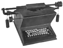 A wooden typewriter with a "QWE-TY" keyboard composed of circular, white keys. The platen rests on top of the device, connected to a bar which extends several inches beyond the sides of the machine. Counterweights are suspended from each end of the bar, to which several wheels and gears are also attached.