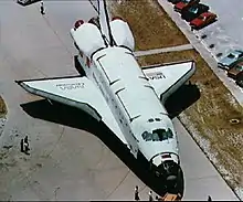 Challenger rolls on its landing gear across a roadway and near parked cars and a few people standing around it.