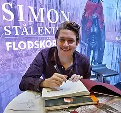 Simon Stålenhag wearing a dark purple patterned shirt with lanyard, sitting at a table in front of a promotional poster of his art, appearing to autograph a book and grinning directly at camera