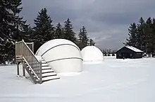 Two white observatory domes in a snow-covered field, wooden stairs are beside one dome and a cabin and trees are in the background