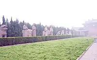 A number of stone statues, mostly of humans and lions, flanking a long paved road that leads to a large building obscured by fog or haze. The statues are separated from the road by a short hedge.