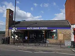 A brown-bricked building with a rectangular, dark blue sign reading "SOUTH EALING STATION" in white letters all under a light blue sky