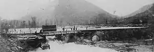 Black and white image of a wide dam made of logs with a rectangular opening in the center, through which logs and water are flowing. The background is mountainous.