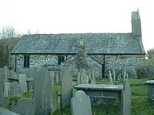 The north side of stone church with a slate roof, seen through a graveyard. There are two windows, a protruding vestry and, at the right, a bellcote