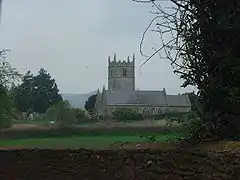 Gray stone building with square tower behind. In the foregound are green fields and bushes.