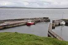 Picture of a small harbour sheltering a red and white fishing boat and a white yacht, with the mainland coast visible in the distance behind