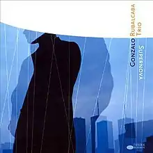 A landscape illustrated in blue with the silhouette of a man in a raincoat and beret tilted to its left front perspective. In the background you see three buildings out of focus. On top a semicircle in white. In the right corner we see the text "Gonzalo Rubalcaba Trio Supernova" on black, white and orange colored font.