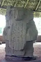 A relief sculpture of a human bust facing forwards, with sculpted cloth hanging over the chest bearing a criss-cross design. The face has a damaged right cheek and two round earspools decorate the ears.