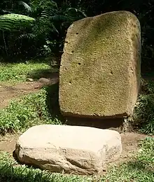 A moss-covered upright stone slab, fallen slightly from vertical. The slab has a number of small cup-like depressions in the surface. In front of it is a horizontal roughly square slab of pale stone with faint designs sculpted around the edges.