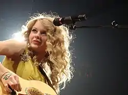 Swift playing a guitar, donning a yellow dress