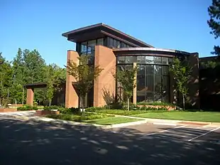 A tan brick building with dark glass walls, including a large curved glass wall facing the viewer. In front of the building are trees and a lawn, and in front of that a driveway and parking spaces.