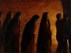 six hooded figures walk rightwards bathed in a dim yellow light from an unseen source