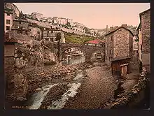 Colorized old postcard showing a low-angle view of the northern part of the valley during low-water periods. In the center, a Roman bridge with 2 arches crosses the river.