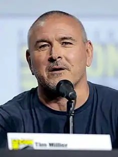 A middle-aged white man in a T-shirt, speaking into a microphone.