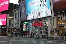 Several retail stores at the ground level of 1540 Broadway, as seen from across Broadway. There is a convenience store, a Disney store, and a Forever 21 from left to right. Above are large billboards.