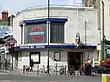 A grey-bricked building with a rectangular, dark blue sign reading "TOOTING BEC STATION" in white letters all under a blue sky with white clouds