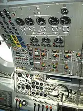 An airliner flight deck control panel with many instruments and switches. A pair of headphones hang from the panel.