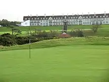 A wide, sprawling golf course. In the background is the Turnberry Hotel, a two-story hotel with white facade and a red roof. This picture was taken in Ayrshire, Scotland.