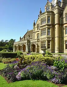 Yellow stone ornate facade of building with lower arched front to the left. In the foreground could flowers in formal garden.