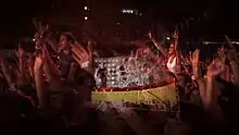 A close-up shot of fans inside a stadium during a concert, many of whom have their arms in the air. A transparent layer on top shows a concert stage with a curved video screen showing black-and-white images of musicians performing, with solid red and yellow colors below.