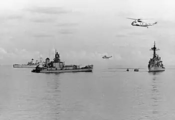 The stern of a destroyer, floating in calm water. The bow has been removed completely. Two intact warships are close by, one to the right of frame, the other behind and partially obscured by the floating stern, and two helicopters are flying overhead.