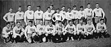 A group of 23 young men arranged in two rows—thirteen standing in back and ten squatting in front—in rugby uniform: white sweaters emblazoned with vertically striped shields, black belts, and light colored shorts.