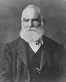 Elderly balding male figure in a dark suit, with a large white beard.