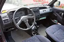  Dashboard of a car with cloth seats, manual gearbox, no airbags, analogue dials, rotary fan and heater controls, a glovebox, hand-cranked windows, and a compact cassette head unit.