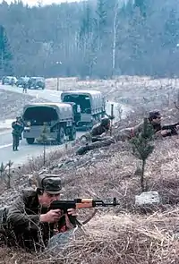 photo of a Slovenian territorial defence member aiming his rifle, with military vehicles in the background