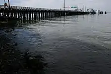 Water is meeting the shore in the foreground of the picture while a pylon based bridge is in the far left corner, which is the ferry dock