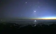 A photograph of the night sky taken from the seashore. Many glimmers of sunlight is on the horizon. There are many stars visible. Venus is at the center, much brighter than any of the stars, and its light can be seen reflected in the ocean.