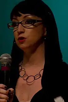 A woman with long black hair, black-framed glasses, red lipstick, black nail polish and hoop earrings and a ring necklace speaks into a mesh-tipped microphone