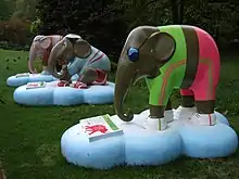 Three brightly coloured elephant statues line up on lush grass by trees in Hyde Park. All three elephants appear to be wearing sporting or athletic gear. Each statue stands on a light blue, cloud-shaped base, with an information plate displaying the artwork details.